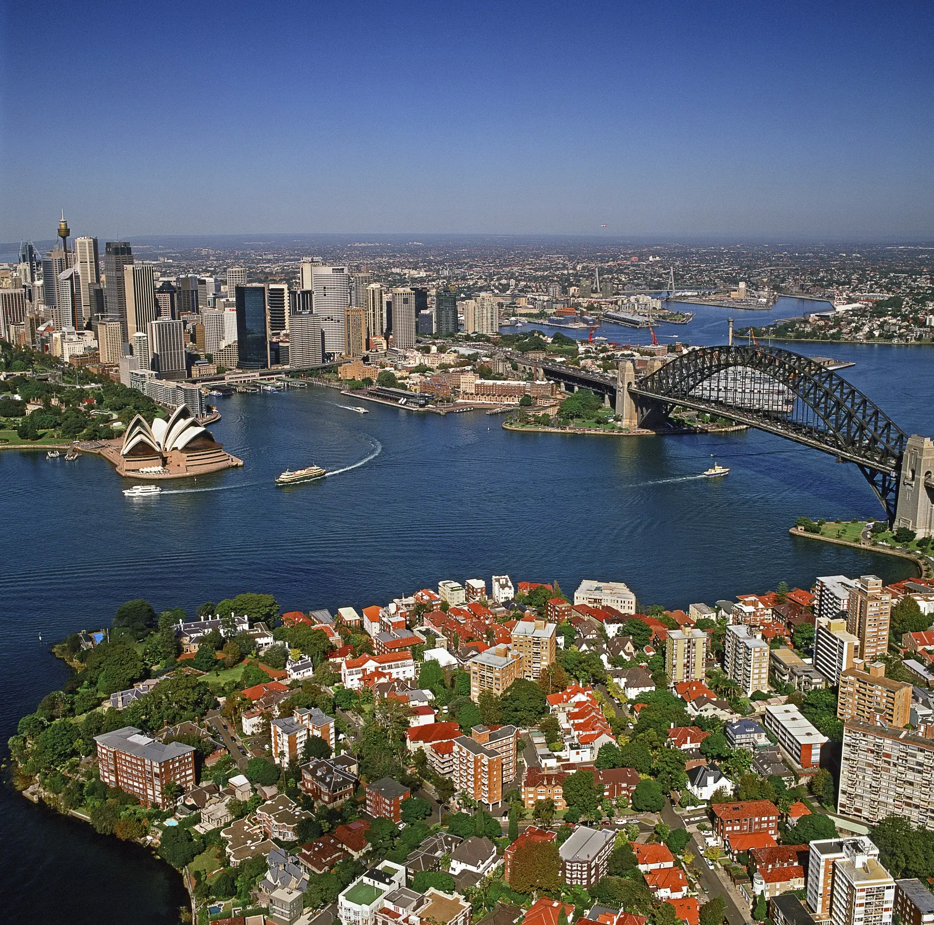 An aerial view captures the scenic skyline of Sydney, Australia during daytime. The iconic Opera House and the Sydney Harbour Bridge are clearly visible, sitting between the bustling city and residential areas with orange tiled roofs. Several ships traverse the blue waters of the bay, and many trees populate the landscape. This photograph provides a compelling visual for travel publications, tourism promotions, architectural studies, or urban planning projects. It conveys themes of exploration, development, and the harmonious blend of nature and city life.