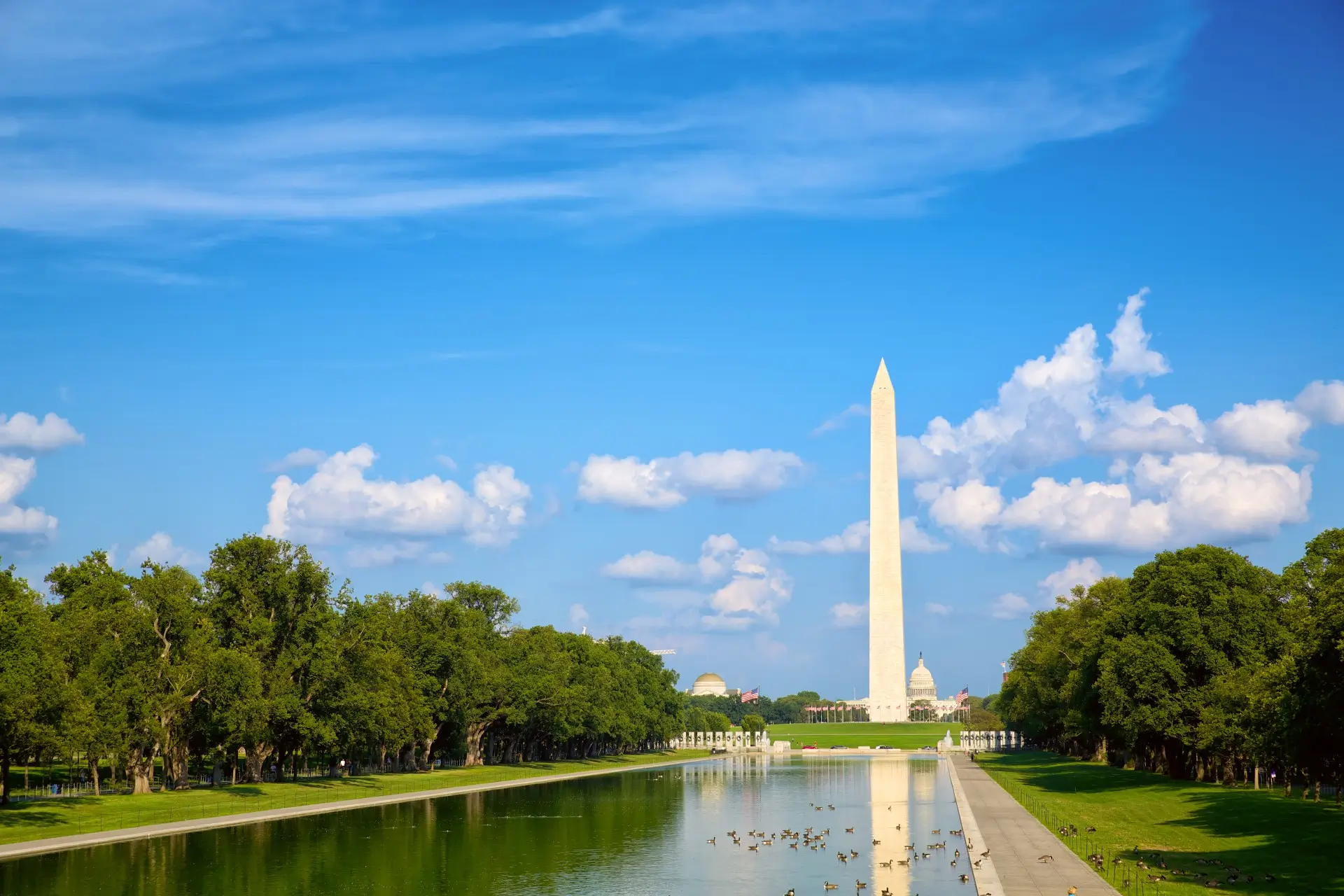 This wide angle shot captures the Washington Monument and reflecting pool during the day. The monument, a tall obelisk, stands in the distance under a blue sky dotted with fluffy white clouds. The reflecting pool runs lengthwise in the foreground, its water reflecting the monument. Flanking either side of the pool are strips of well-manicured green lawn, interspersed with the lush green canopies of trees. A few people are visible walking and enjoying the sunny weather. The scene is peaceful, still, and could be used for projects related to travel, monuments, landmarks, recreation, or American history.